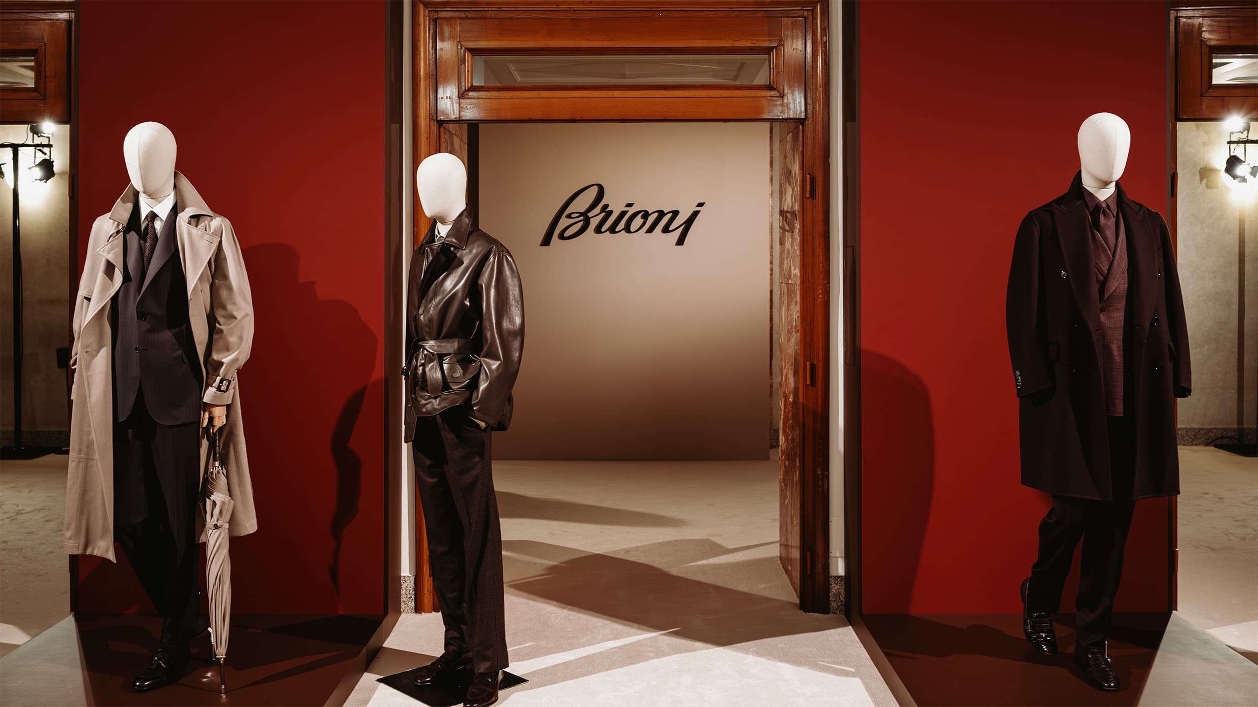 Three mannequins displaying Brioni luxury menswear with coats and suits against red walls in an elegant showroom.