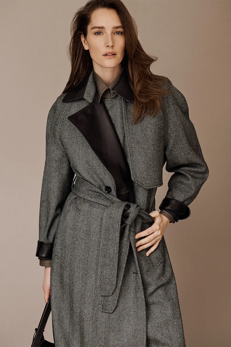 Woman wearing a Brioni gray herringbone coat with black leather collar and cuffs against a beige background