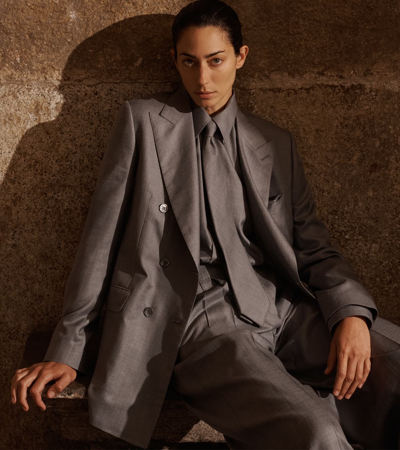 Woman in Brioni gray tailored suit sitting against textured brown wall, looking directly at camera with slicked-back dark hair.