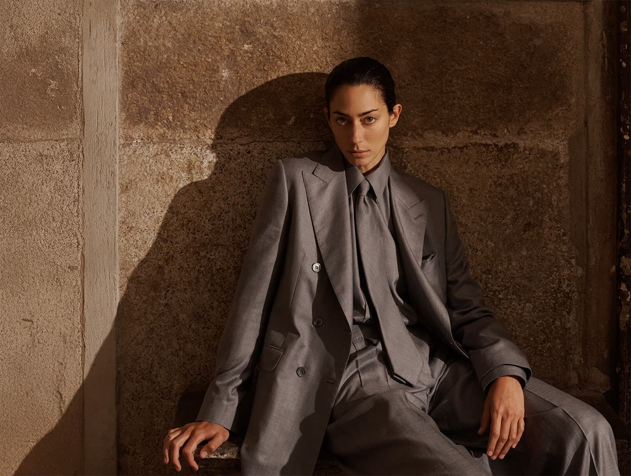 Woman in Brioni gray tailored suit sitting against textured brown wall, looking directly at camera with serious expression.