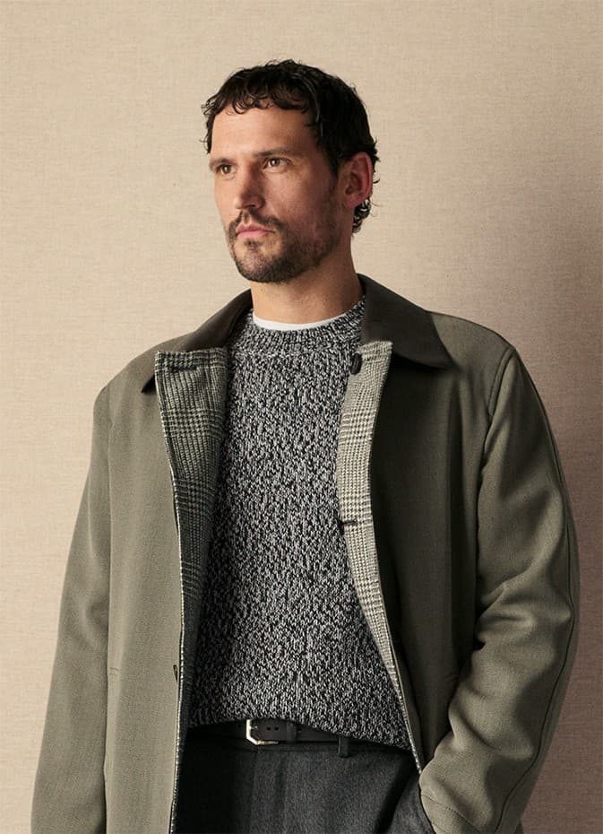 Man with beard wearing Brioni gray textured sweater and olive green jacket with black collar against beige background