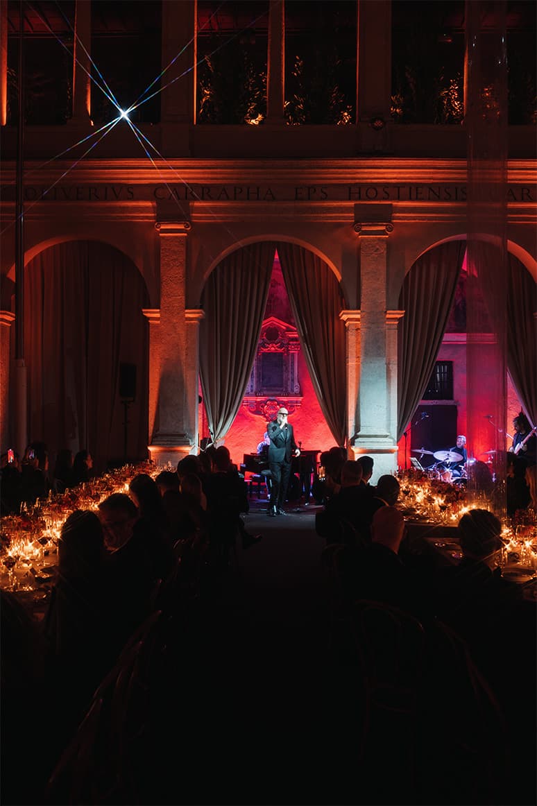 Elegant Brioni gala dinner with performer on stage in historic venue illuminated by red lighting and candlelit tables.