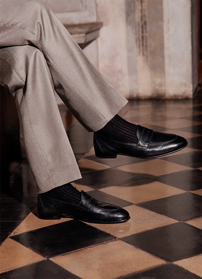Person wearing Brioni black leather loafers and beige wide-leg trousers standing on checkered tile floor in vintage interior.
