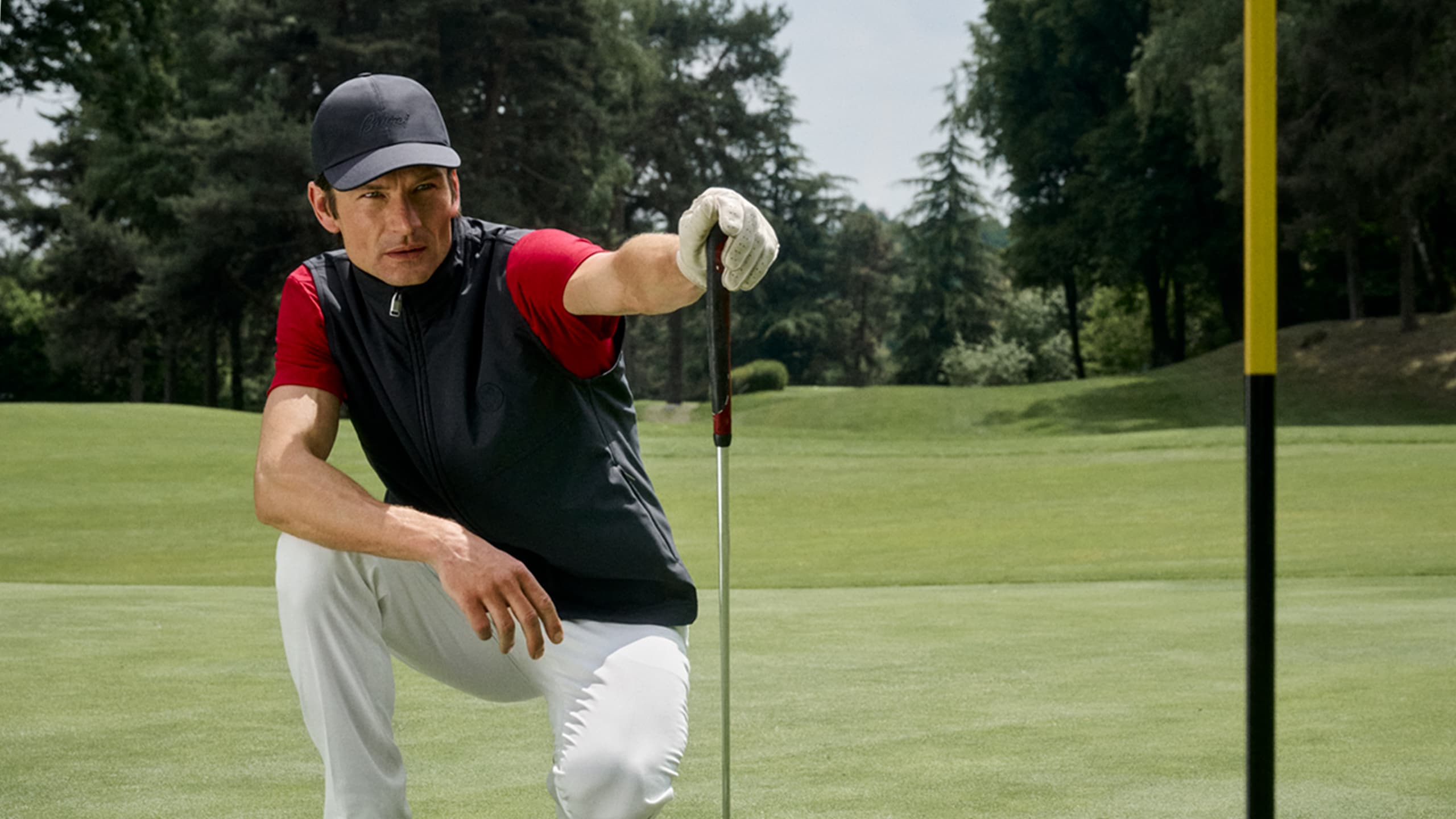Golfer in black cap and vest with red sleeves crouching on green to analyze putt, surrounded by trees.