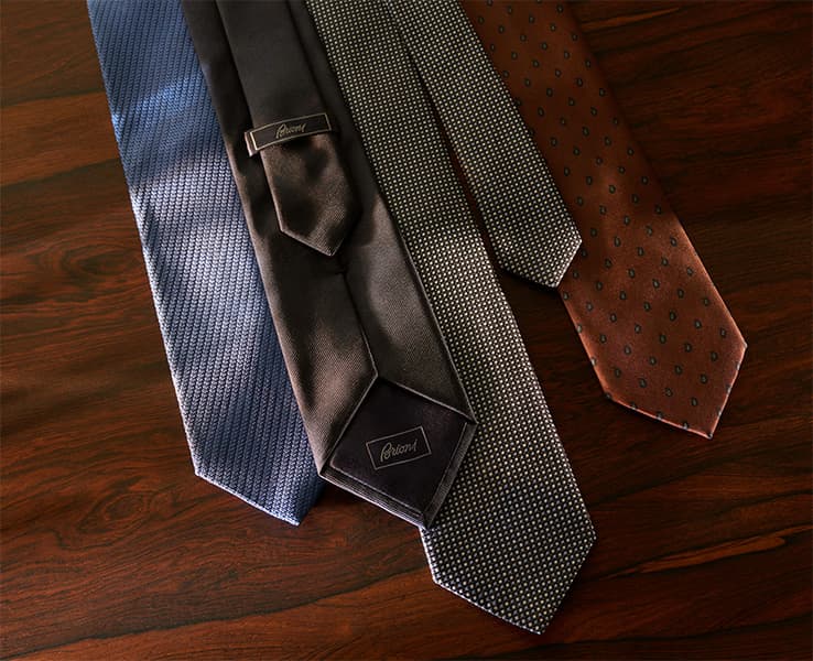 Collection of four elegant Brioni men's ties in blue, black, patterned gray, and rust brown displayed on wooden surface.