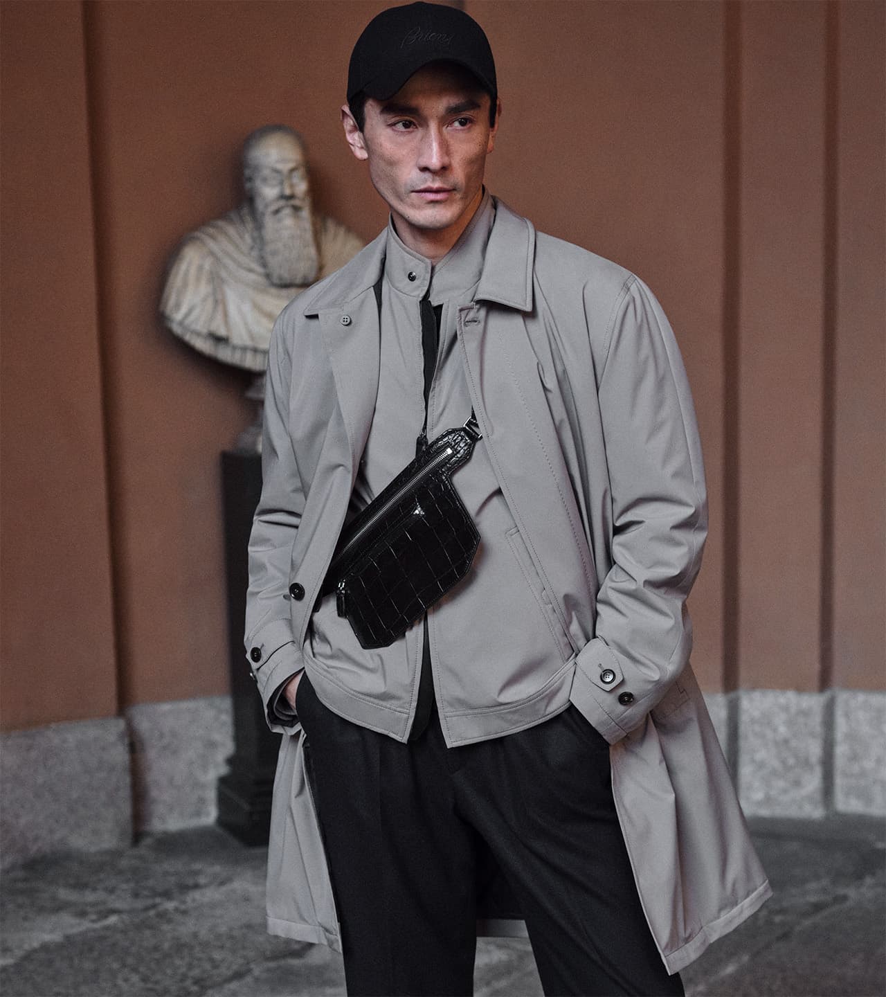 Person in beige jacket and black cap wearing crossbody bag, standing in front of classical bust sculpture.