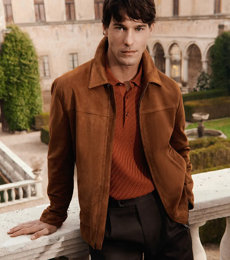 Man in brown Brioni suede jacket and rust-colored polo shirt standing on terrace with historic building and manicured gardens behind him.