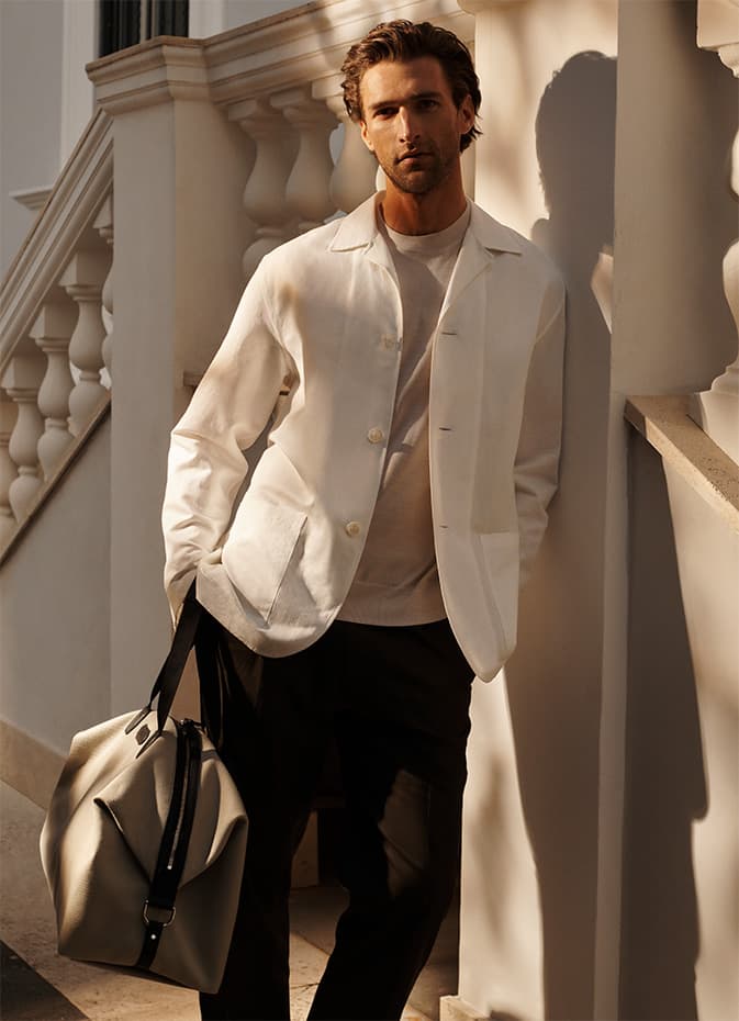 Man in white Brioni blazer and black pants holding beige bag, standing by white columns with warm sunlight casting shadows.