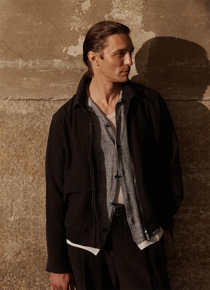 Profile of person in black Brioni leather jacket and gray shirt against textured beige wall, casting dramatic shadow in warm lighting.