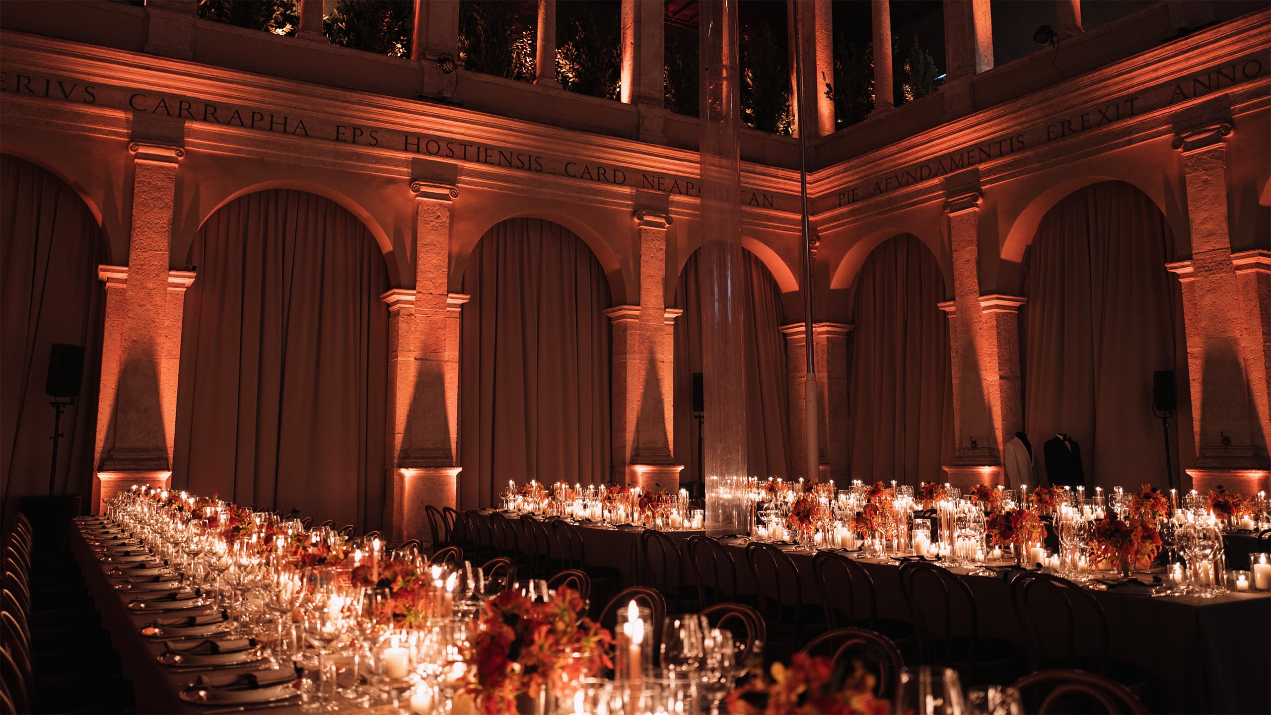 Elegant banquet tables with candles and floral arrangements in a historic hall with arched columns bathed in amber light.