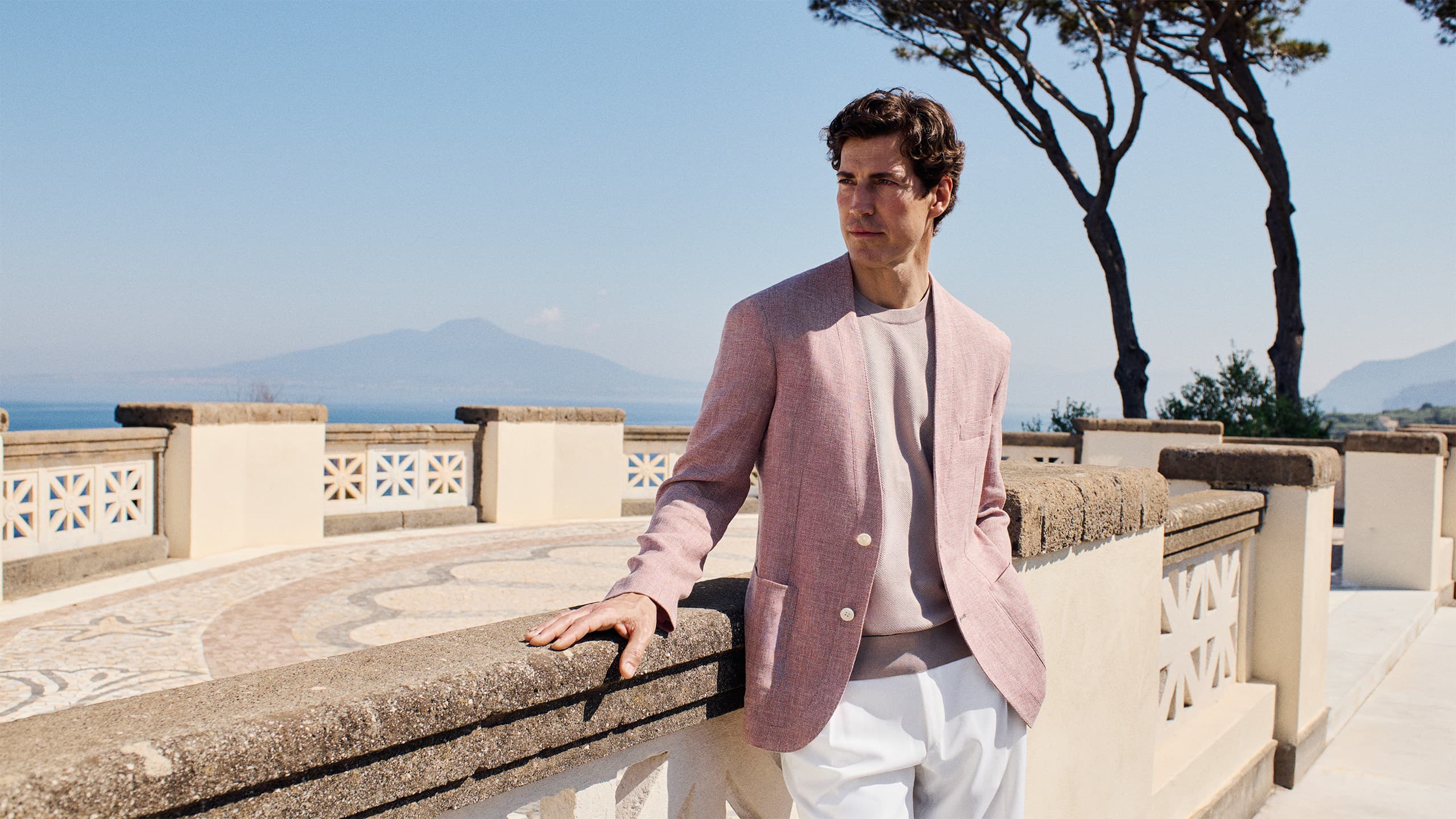 Man in Brioni pink linen blazer standing on Mediterranean terrace with coastal mountain views and pine trees under clear blue sky.