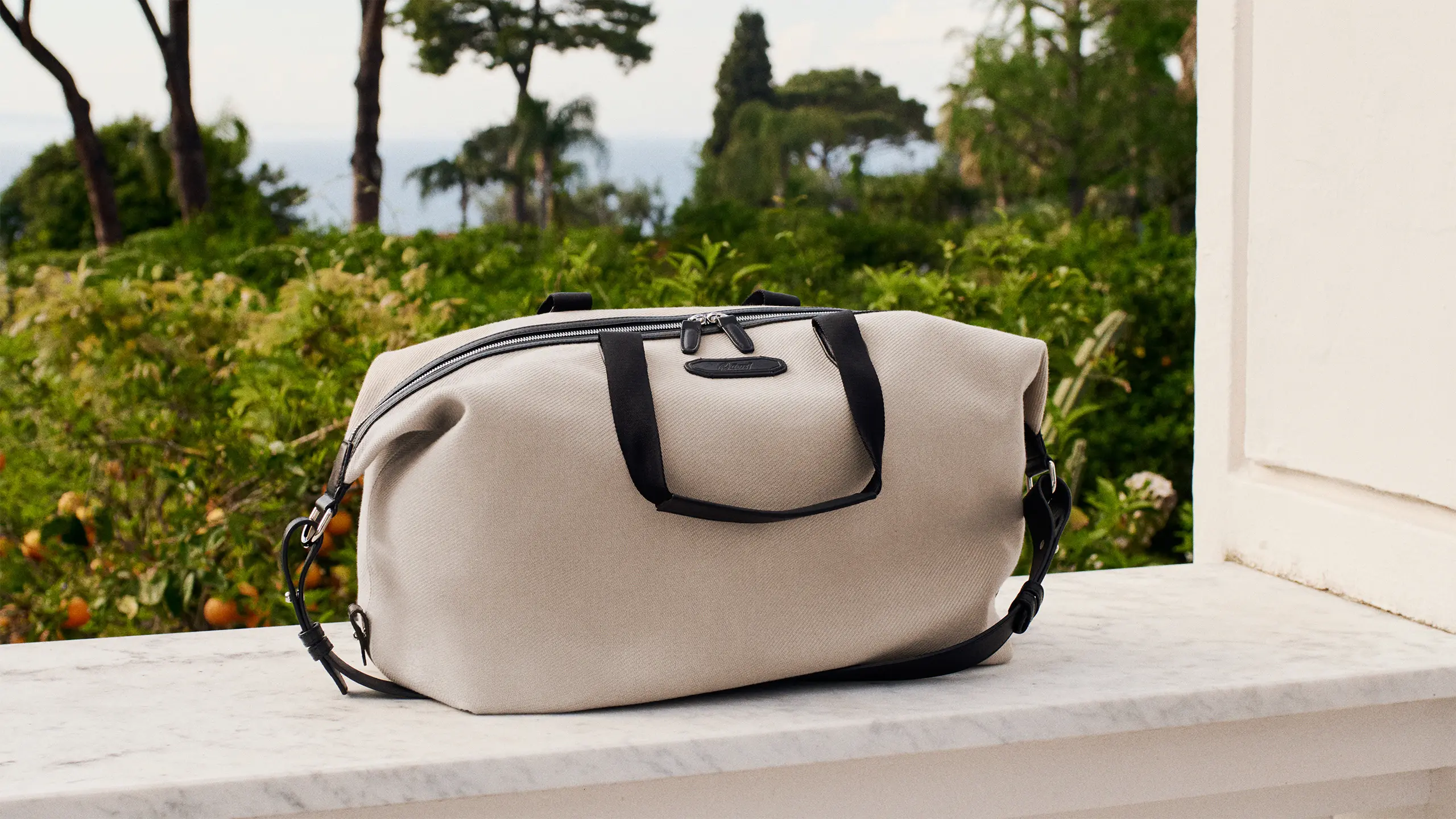 Beige duffel Brioni bag with black handles and strap on white marble surface with lush green garden and ocean view in background.