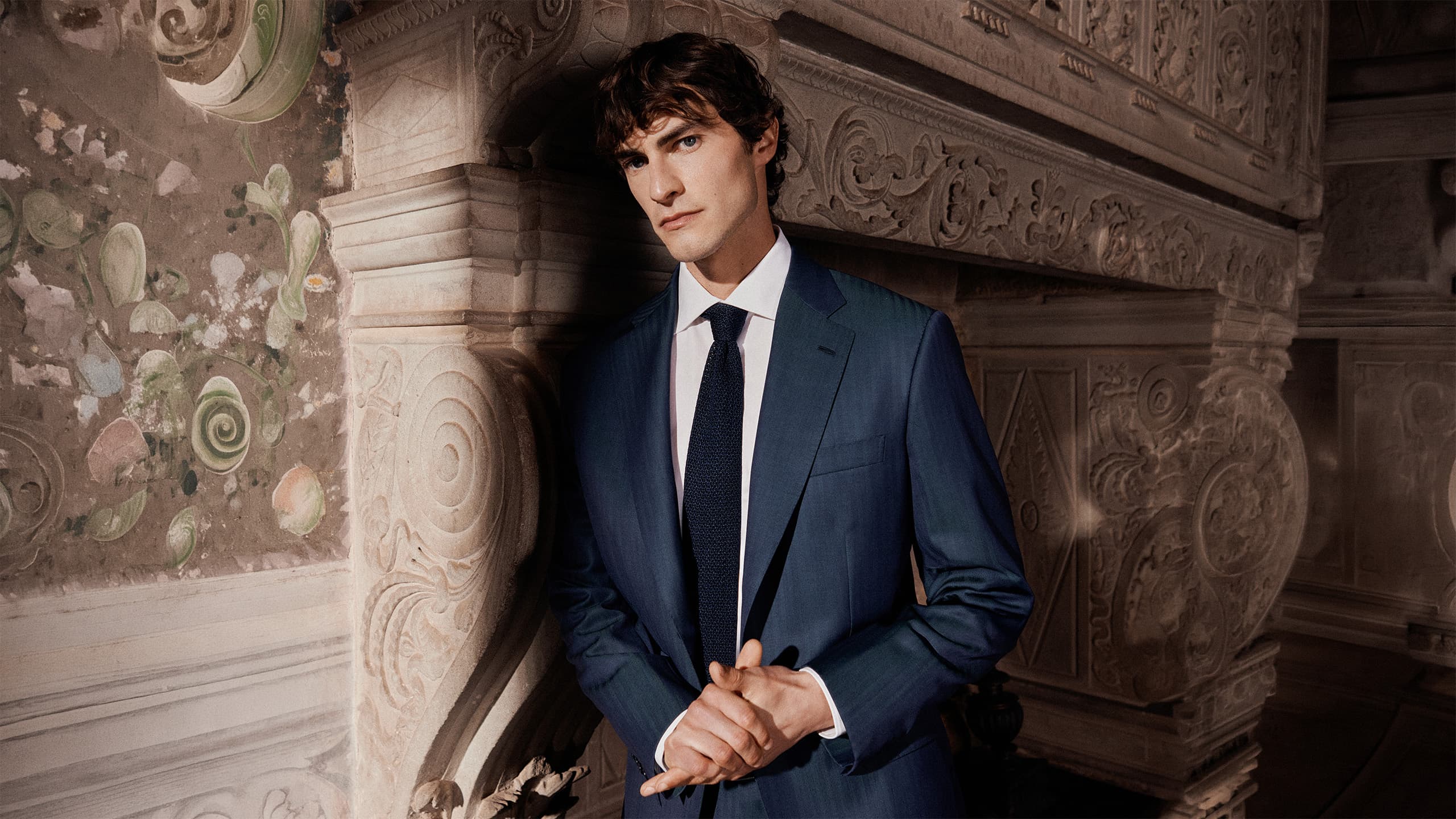 Man in Brioni Iride navy blue suit with white shirt and dark tie standing against ornate stone architecture with decorative carvings.