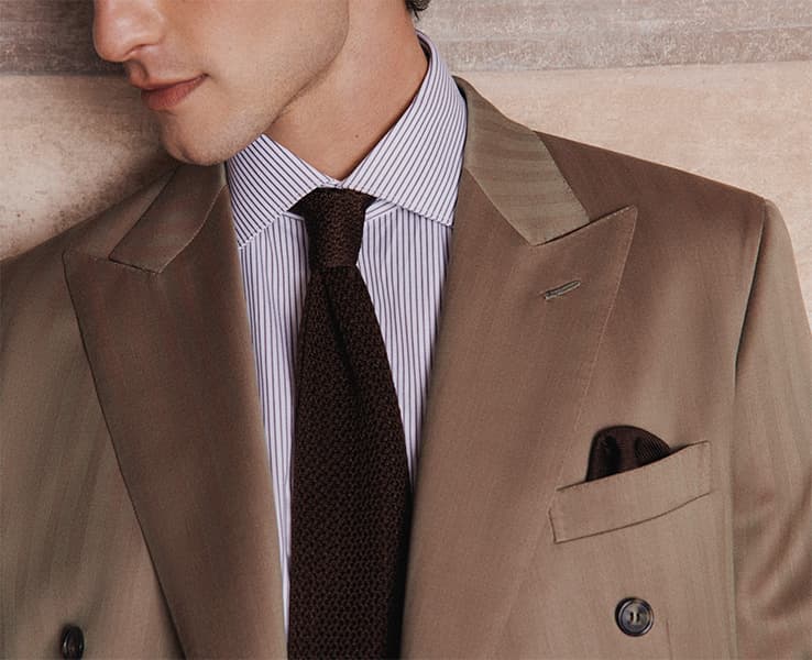 Man wearing Brioni beige double-breasted Iride suit jacket with blue striped shirt, navy blue knit tie, and pocket square against beige wall.