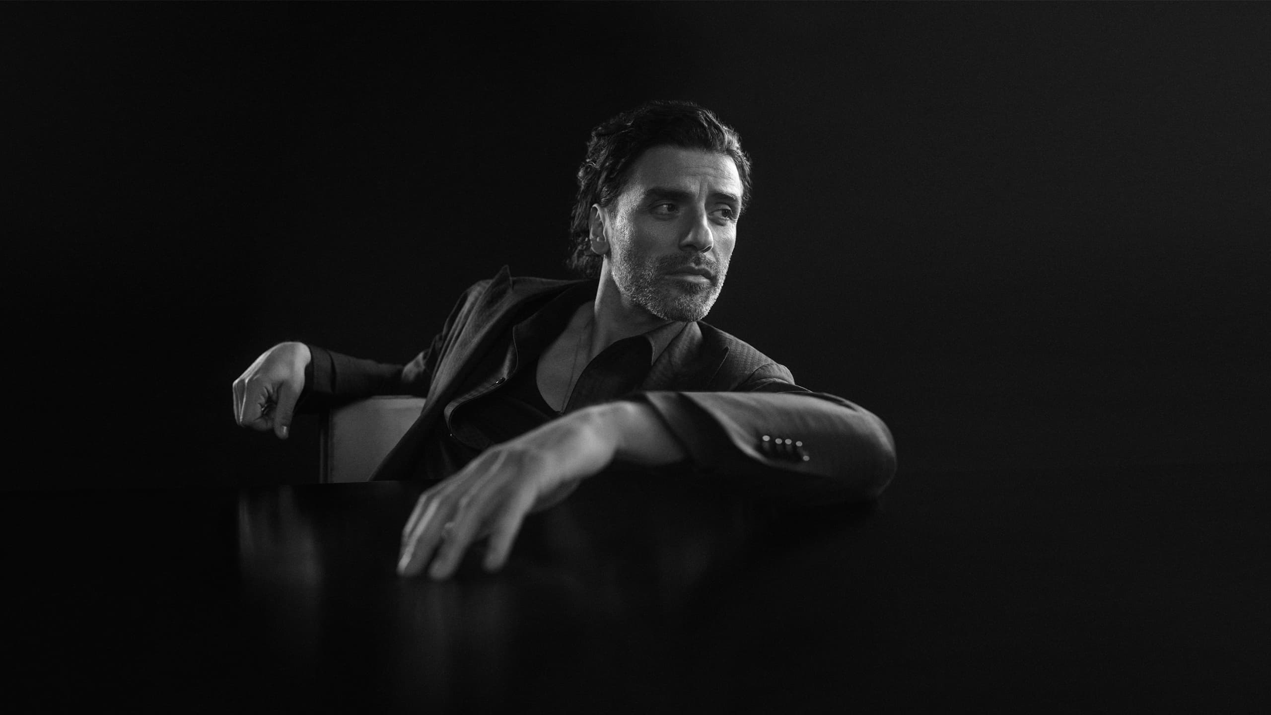 A black and white portrait of Oscar Isaac in a formal Brioni suit attire seated in a chair against a dark background.