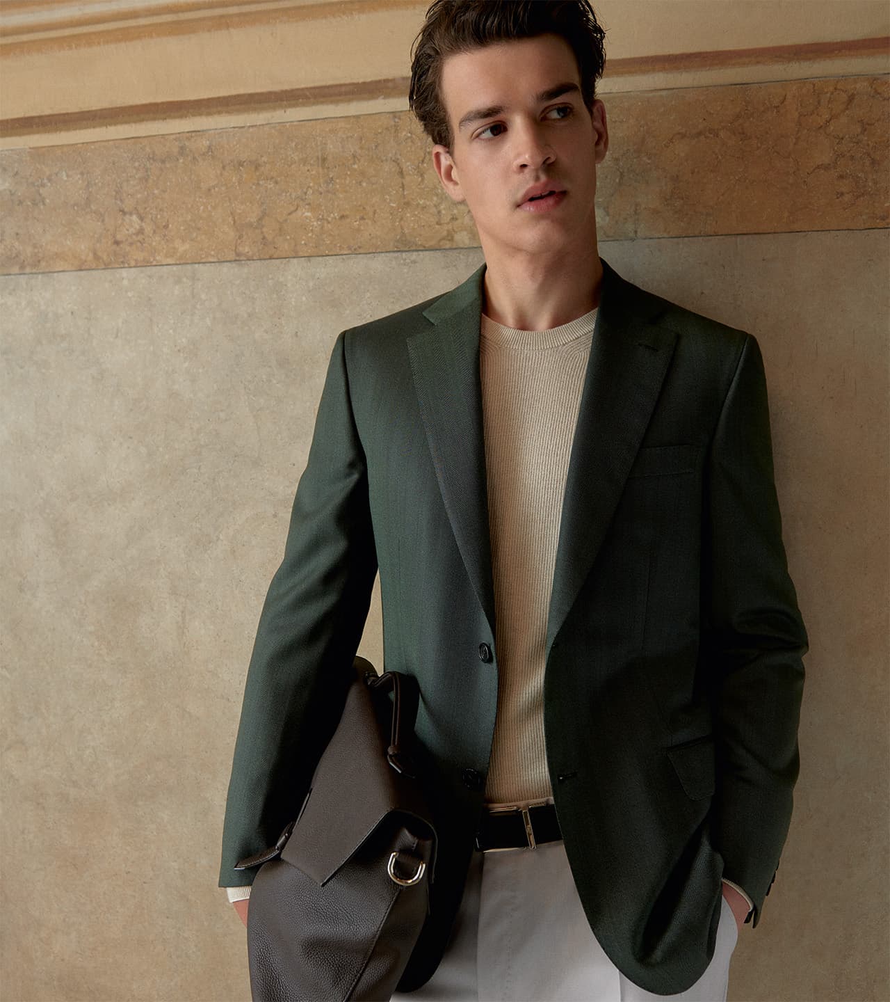 Young man in Brioni dark green blazer and beige sweater holding leather bag against textured stone wall background.