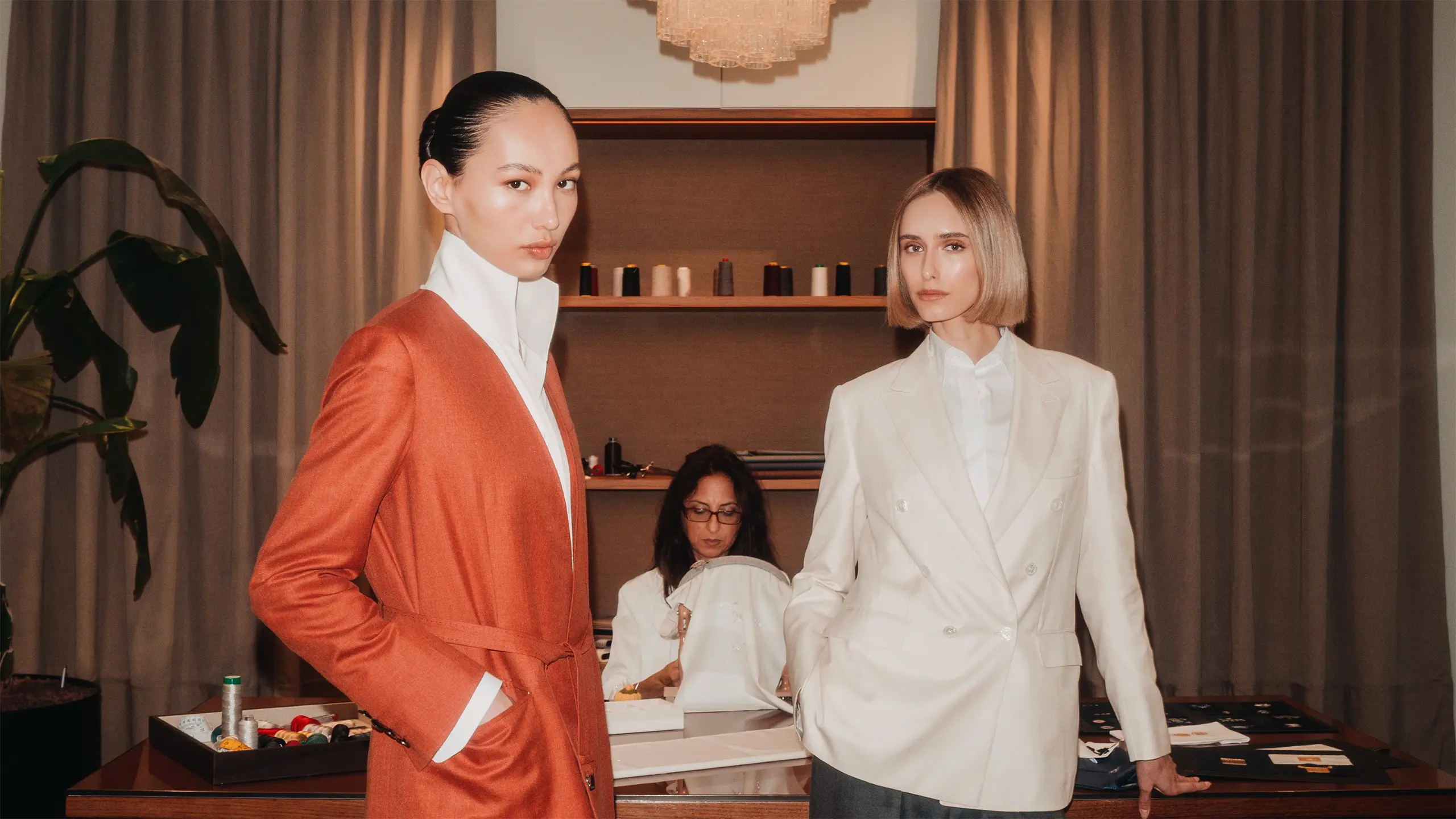Two women in Brioni tailored blazers, one orange and one white, stand in a modern retail space with neutral tones and shelving.