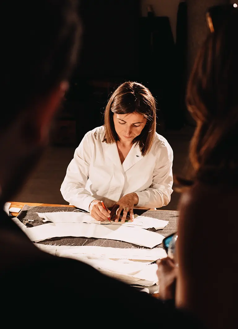 A Tailor drawing on papers at a dimly lit table, surrounded by others in a workshop setting.