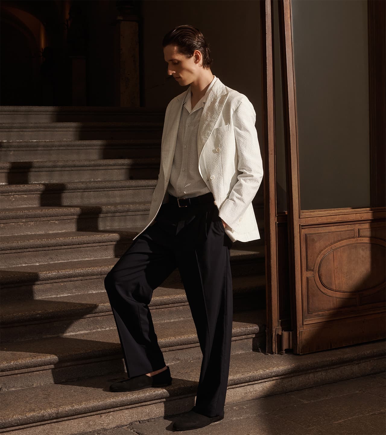 Man in Brioni white blazer and black trousers standing on stone staircase in warm sunlight near wooden door panel.
