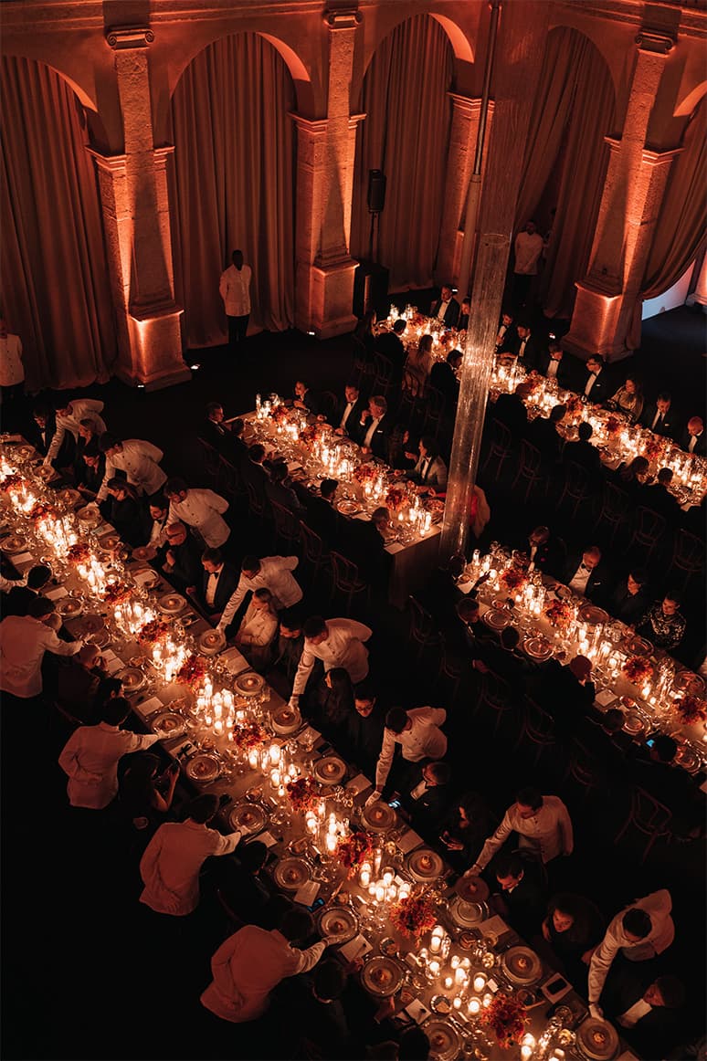 Elegant Brioni formal dinner in grand hall with arched columns, candlelit tables, and servers in white jackets attending guests.