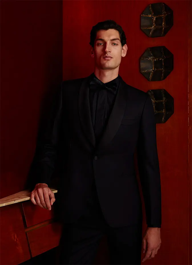 Person in elegant Brioni black tuxedo against rich red wall with decorative octagonal wall hangings.