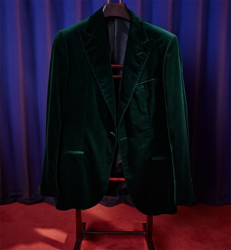 Elegant Brioni emerald green velvet blazer displayed on wooden hanger against purple curtain backdrop and red carpet.