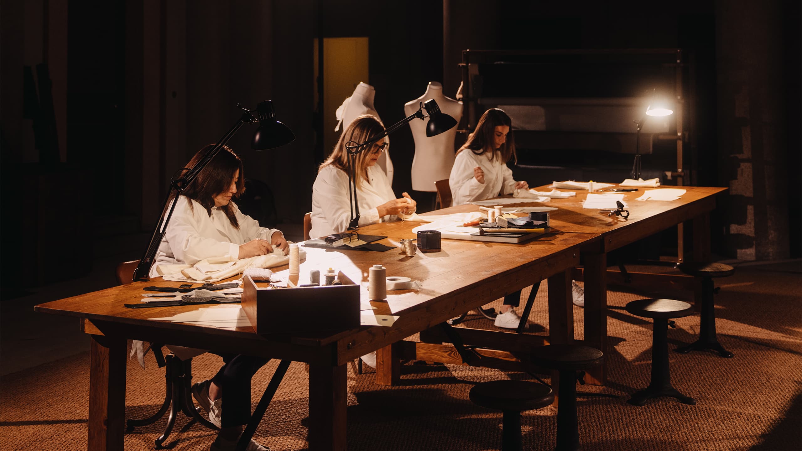 Three tailors working at a long wooden table with sewing materials and desk lamps in a dimly lit studio.