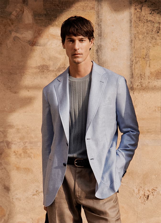 Young man in light blue Soffio jacket and gray crewneck sweater against weathered beige wall with dappled sunlight shadows.