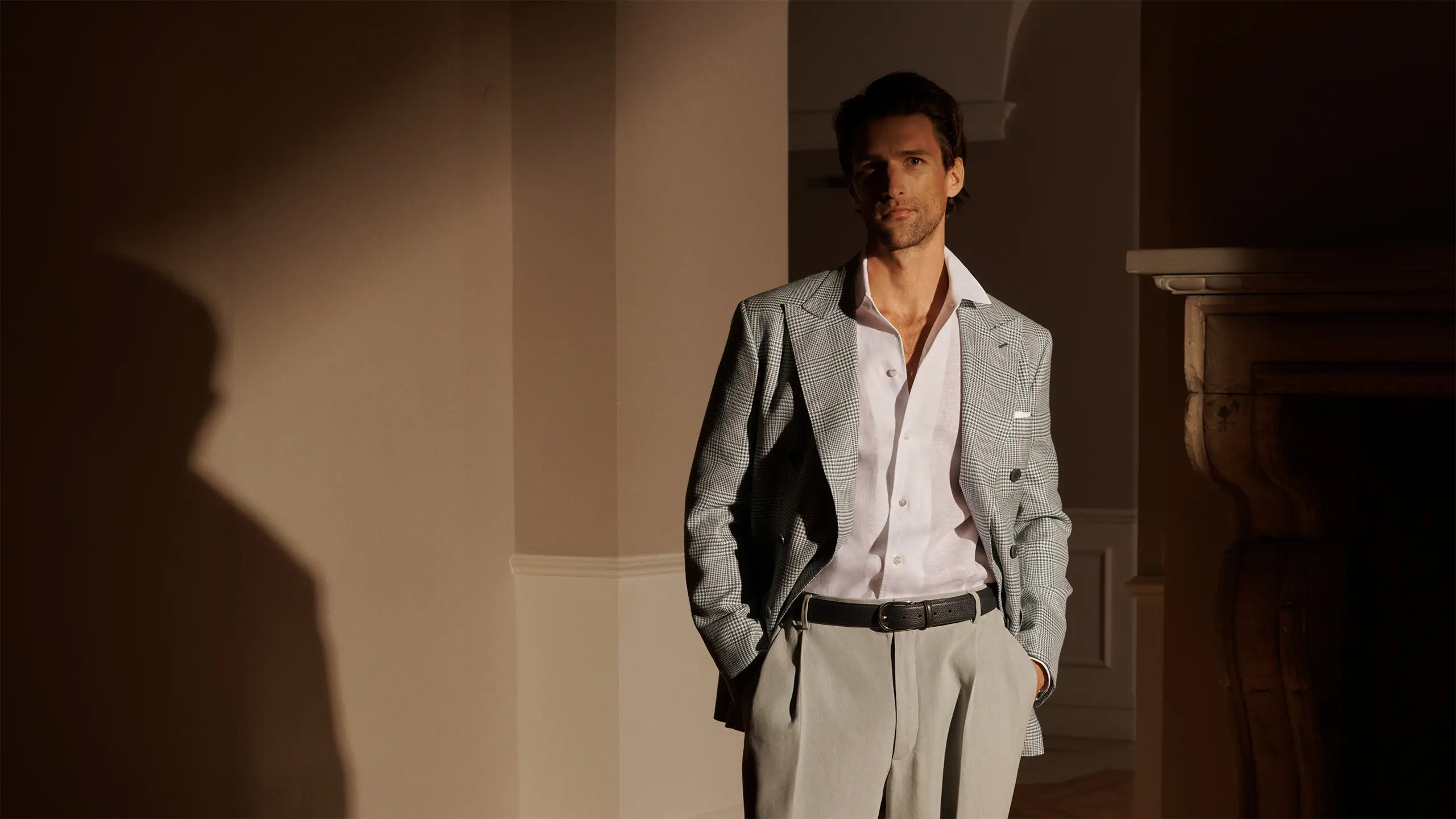 Man in petroleum blue blazer and white shirt standing in warm-lit archway with beige walls and dramatic shadows.