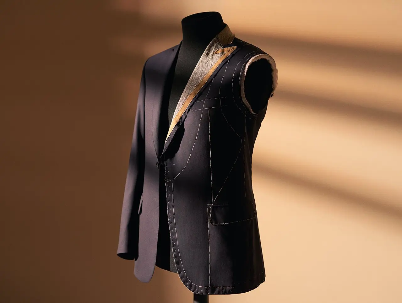 Navy blue tailored Brioni suit jacket on a mannequin showing basting stitches and construction details against warm backdrop.