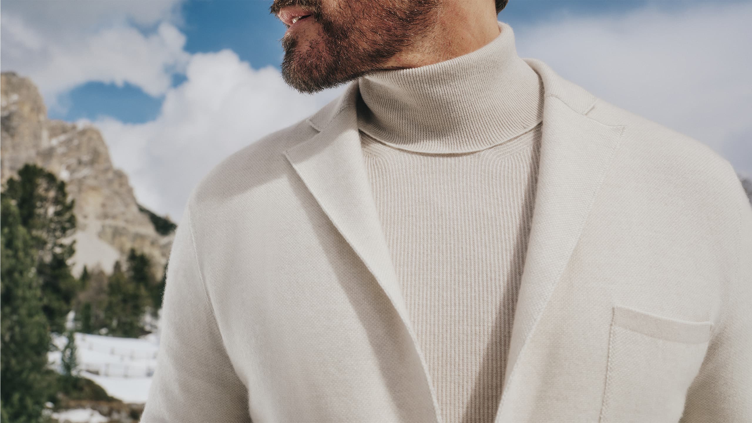 Person wearing Brioni cream turtleneck and blazer against snowy mountain backdrop with blue sky and evergreen trees.