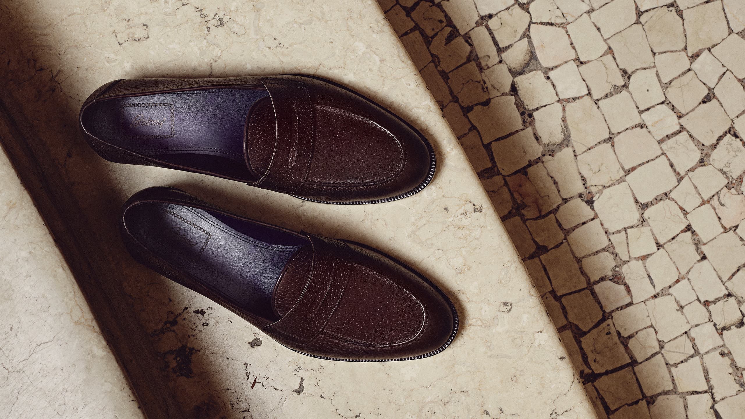 Elegant dark brown leather APPIA loafers on a cream marble surface beside a patterned stone floor.