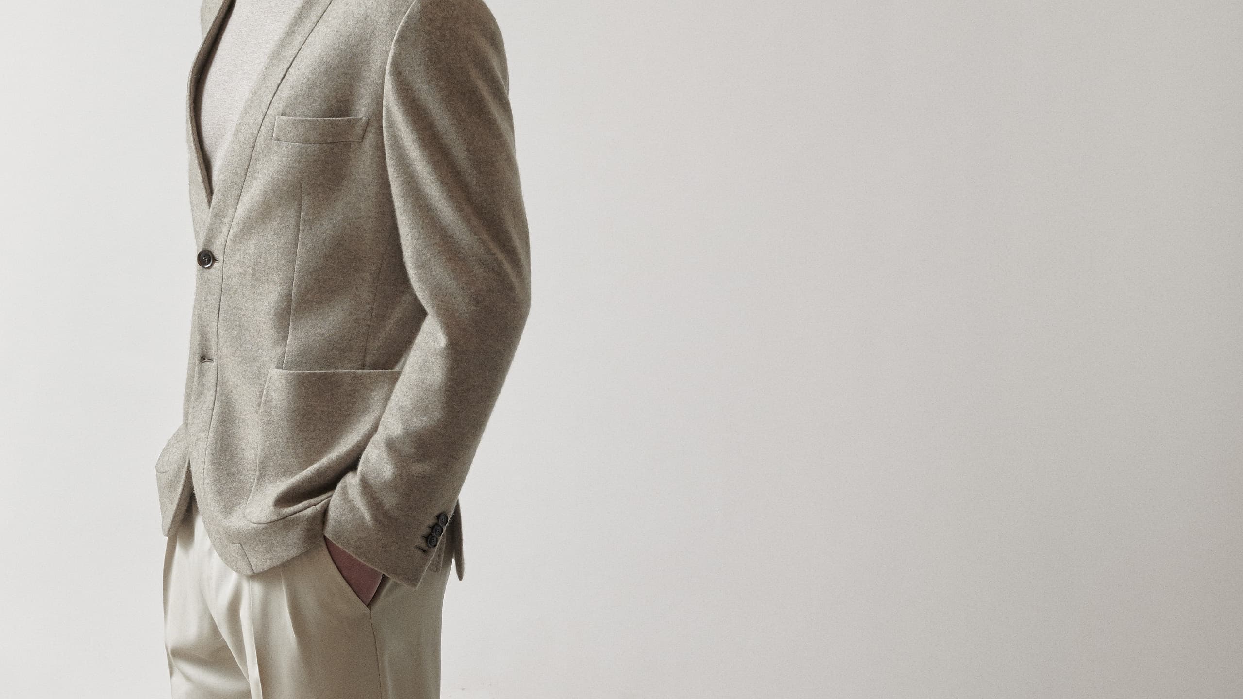Person wearing a tailored beige Brioni blazer with cream trousers, standing with hand in pocket against neutral background.