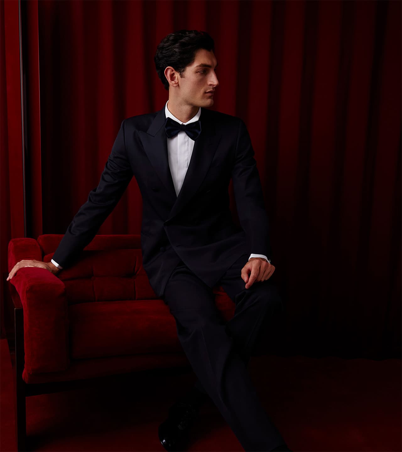 Person in Brioni black tuxedo and bow tie seated on red velvet chair against rich red curtain backdrop.