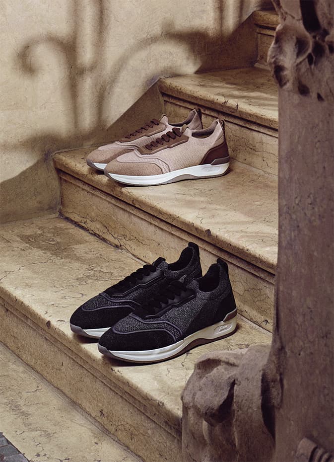 Two stylish Brioni sneakers displayed on stone steps - beige pair on upper step, black pair below with white soles.