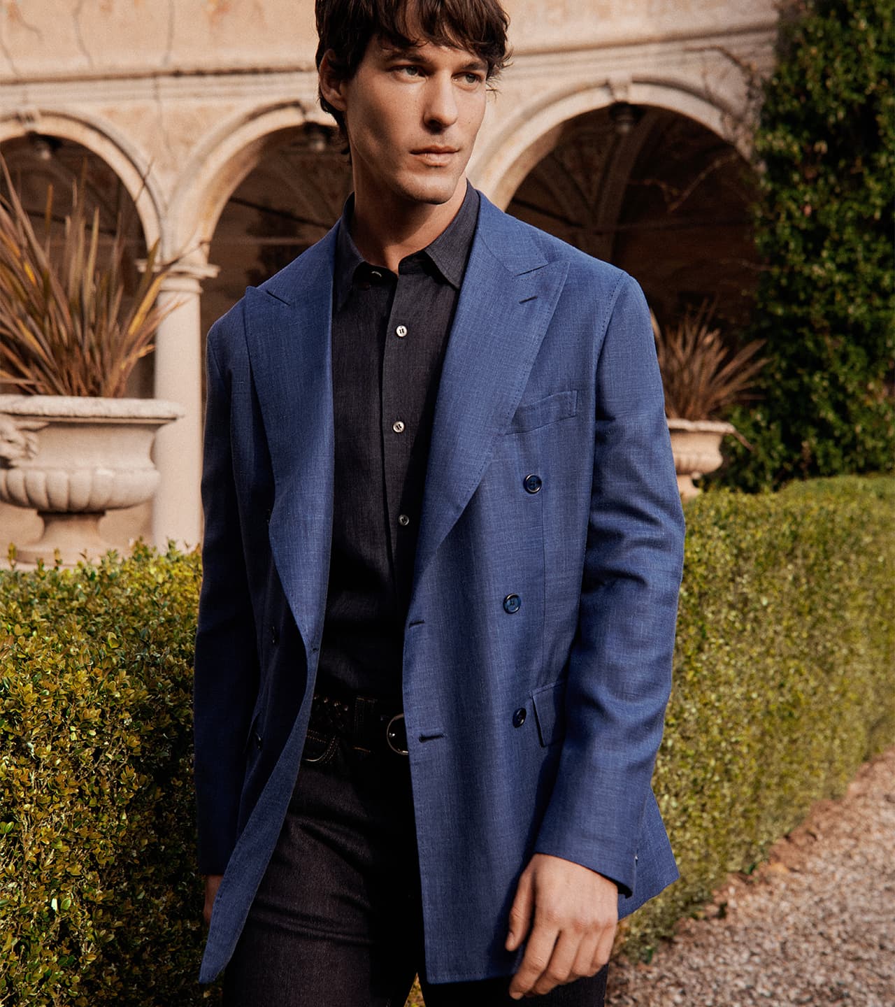 Man in blue Brioni blazer and black shirt standing in garden with stone archway and greenery in background.