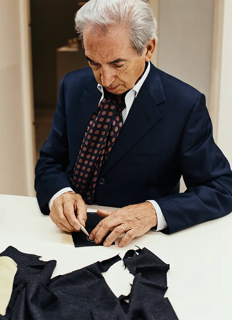Un anziano sarto in abito blu navy con cravatta a fantasia lavora meticolosamente su pezzi di tessuto scuro a un tavolo bianco.