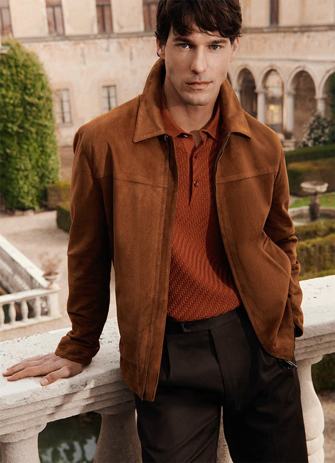Man in Brioni brown suede jacket and rust polo shirt standing by stone balustrade with classical building and garden in background.