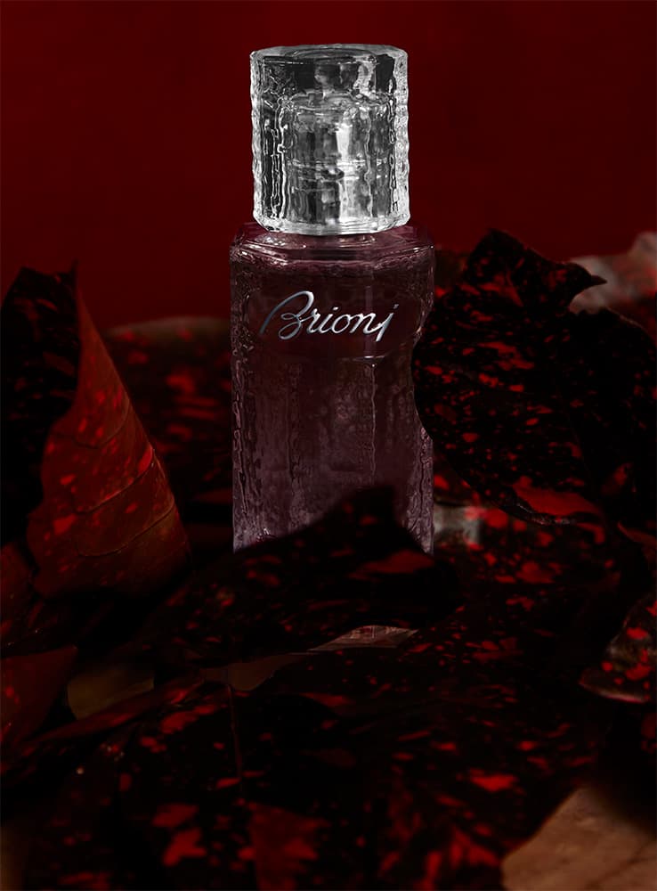 Brioni perfume bottle with crystal cap against deep red background, surrounded by dark textured elements.
