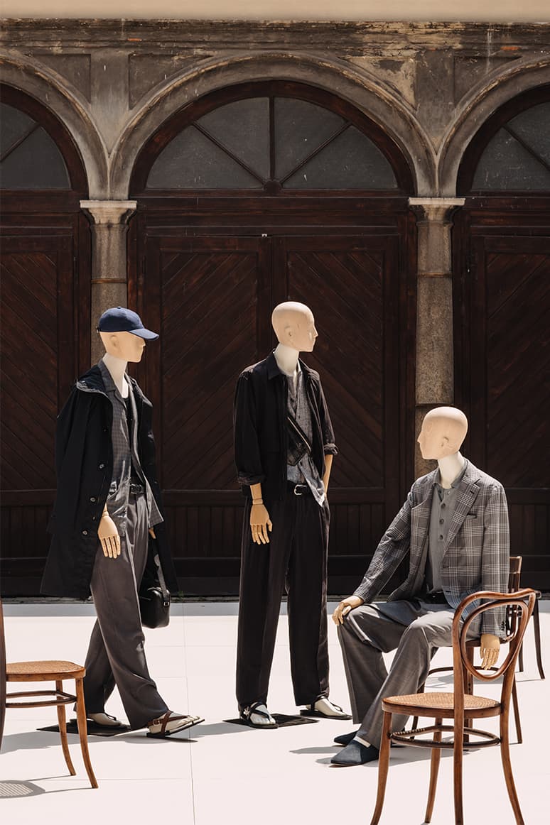 Three mannequins displaying Brioni's ss26 collection in dark suits and coats against an ornate wooden archway backdrop.