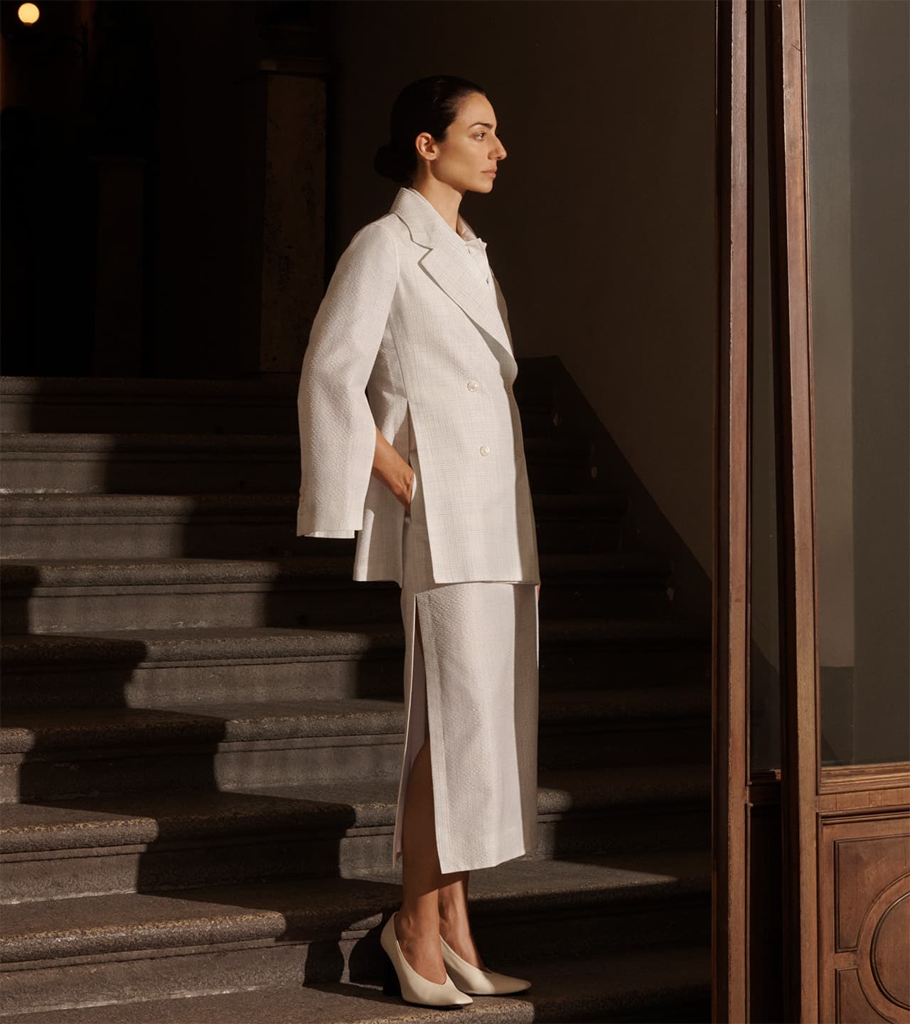 Woman in Brioni white tailored suit standing in profile on stone steps near wooden door, illuminated by warm natural light.