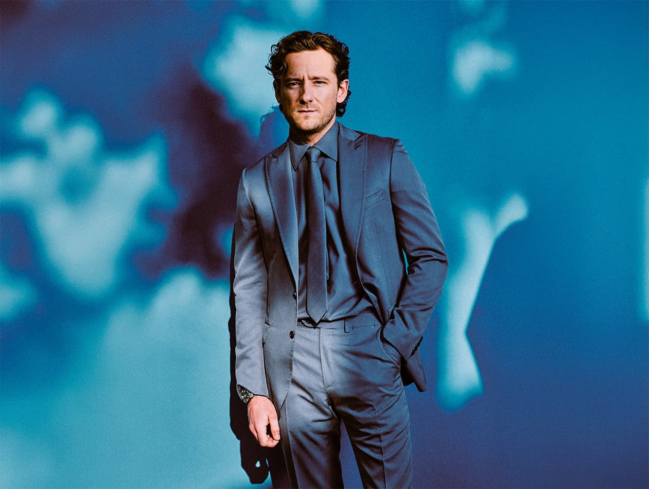 A person in a Brioni navy blue suit and tie standing against a dreamy blue background with abstract light patterns.