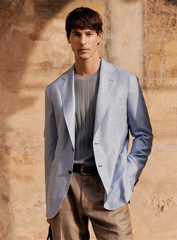 Young man in Brioni light blue blazer and gray sweater standing against weathered beige stone wall with hands in pockets.